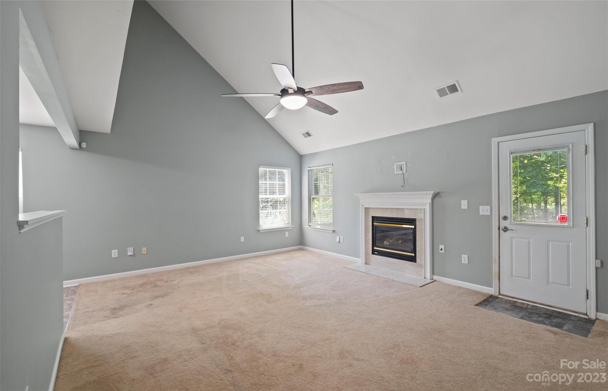 Empty living room with vaulted ceiling, beige carpet, ceiling fan, and gas fireplace.