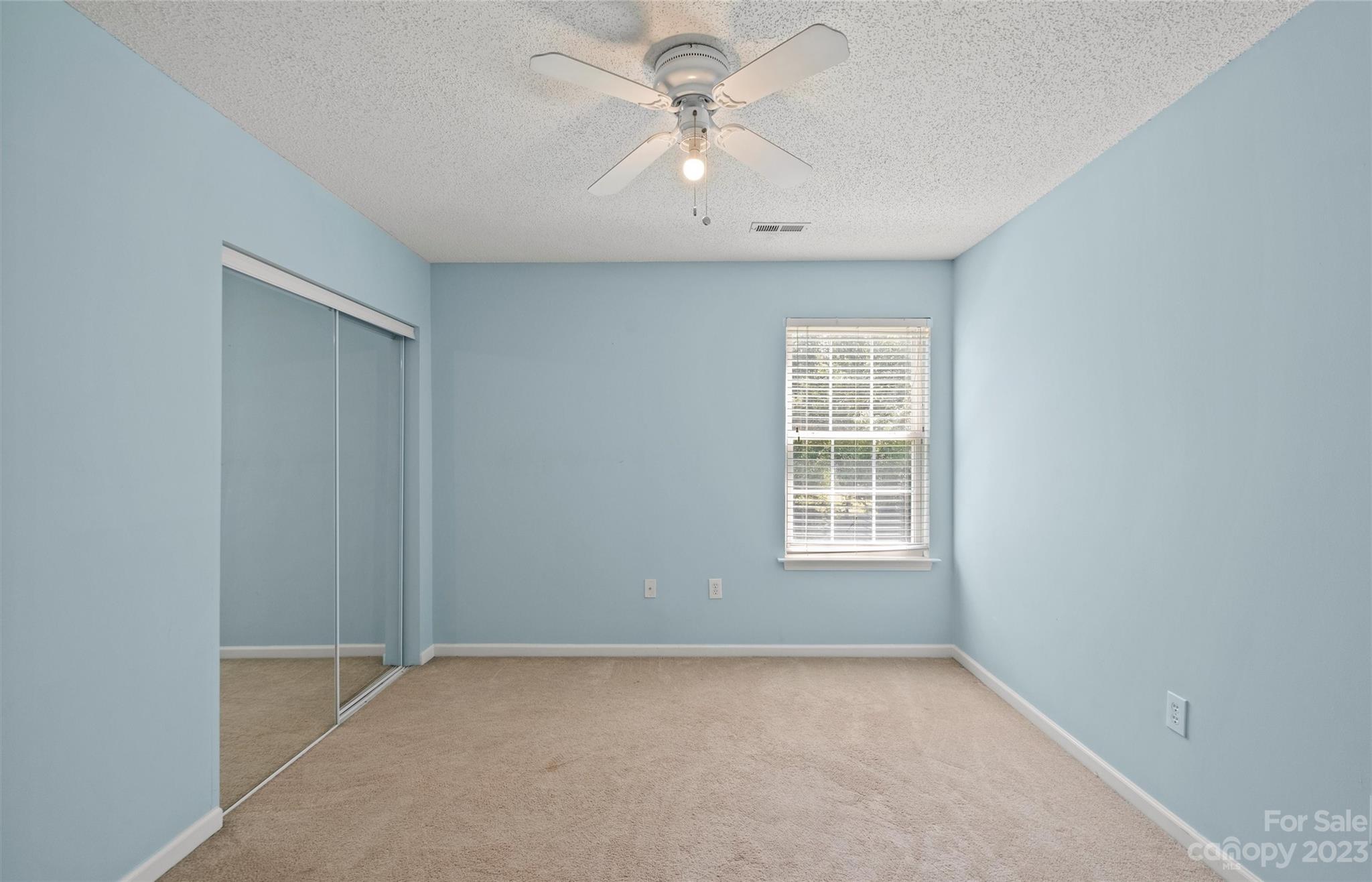 Empty bedroom with blue walls, beige carpet, mirrored closet doors, and a ceiling fan.