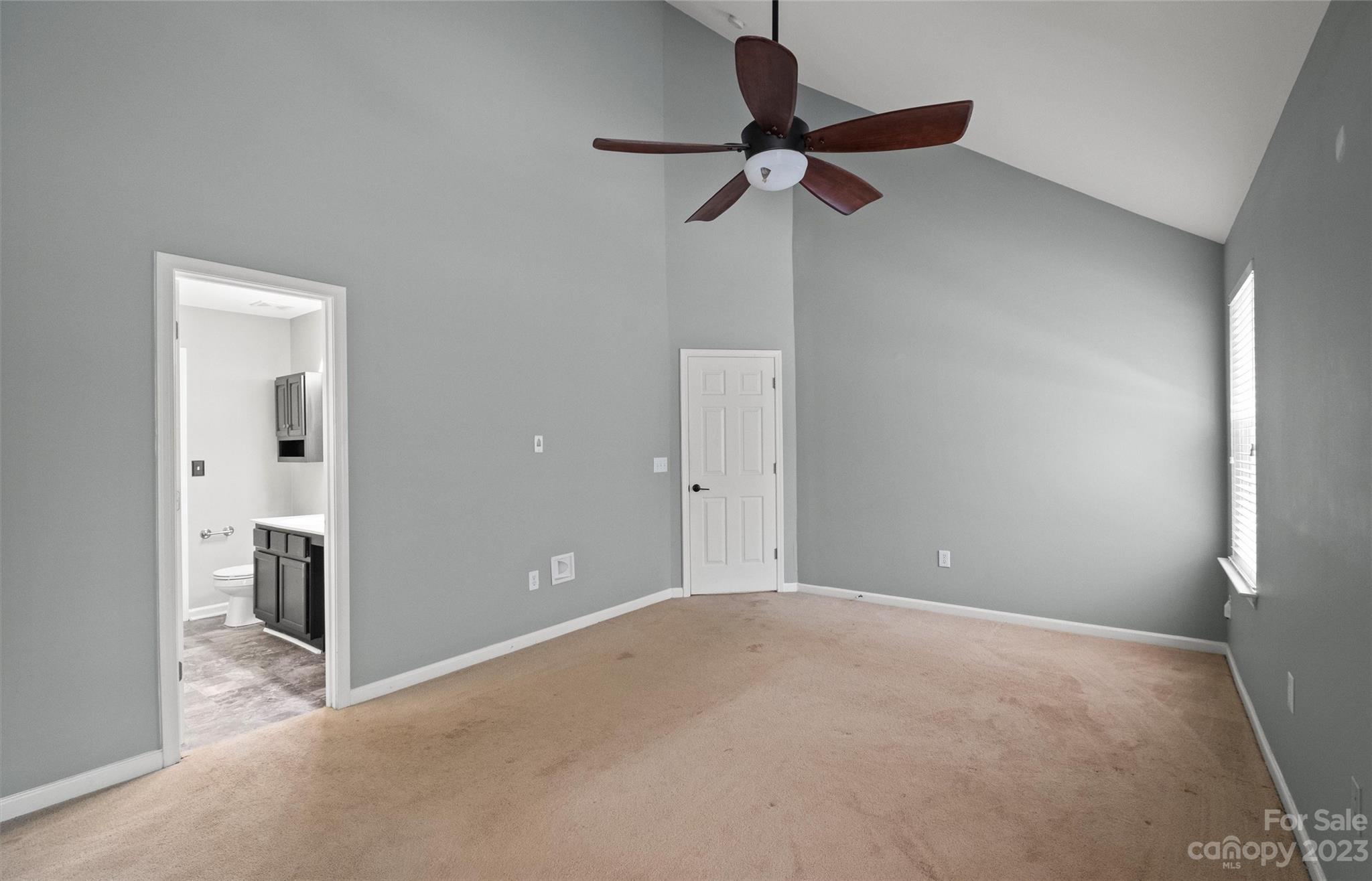Vacant bedroom with a high ceiling, dark wood ceiling fan, beige carpet, and doorway to an adjoining bathroom.