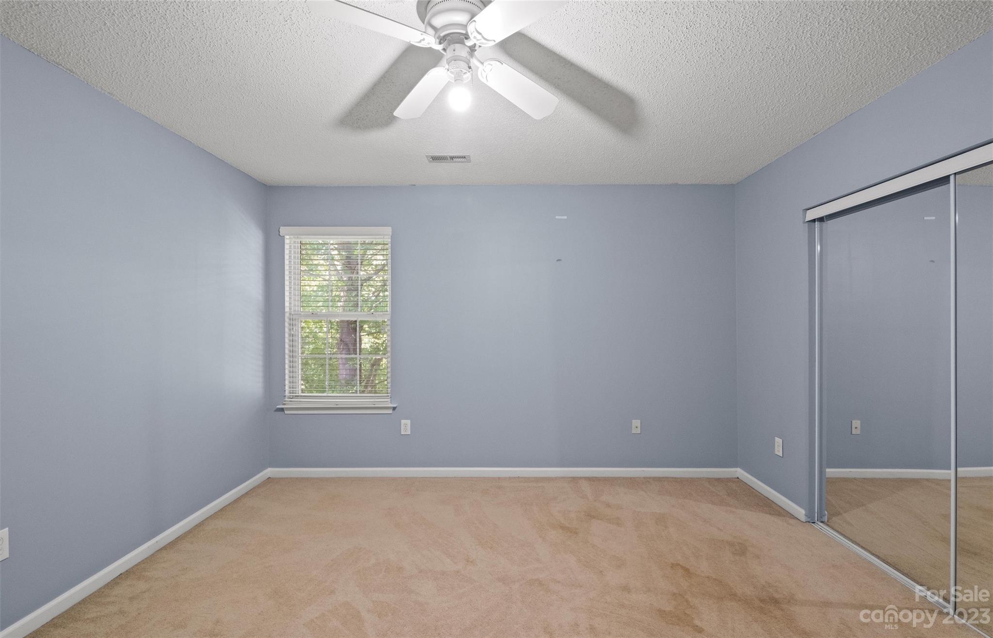 Empty bedroom with beige carpet, mirrored closet doors, and a ceiling fan.