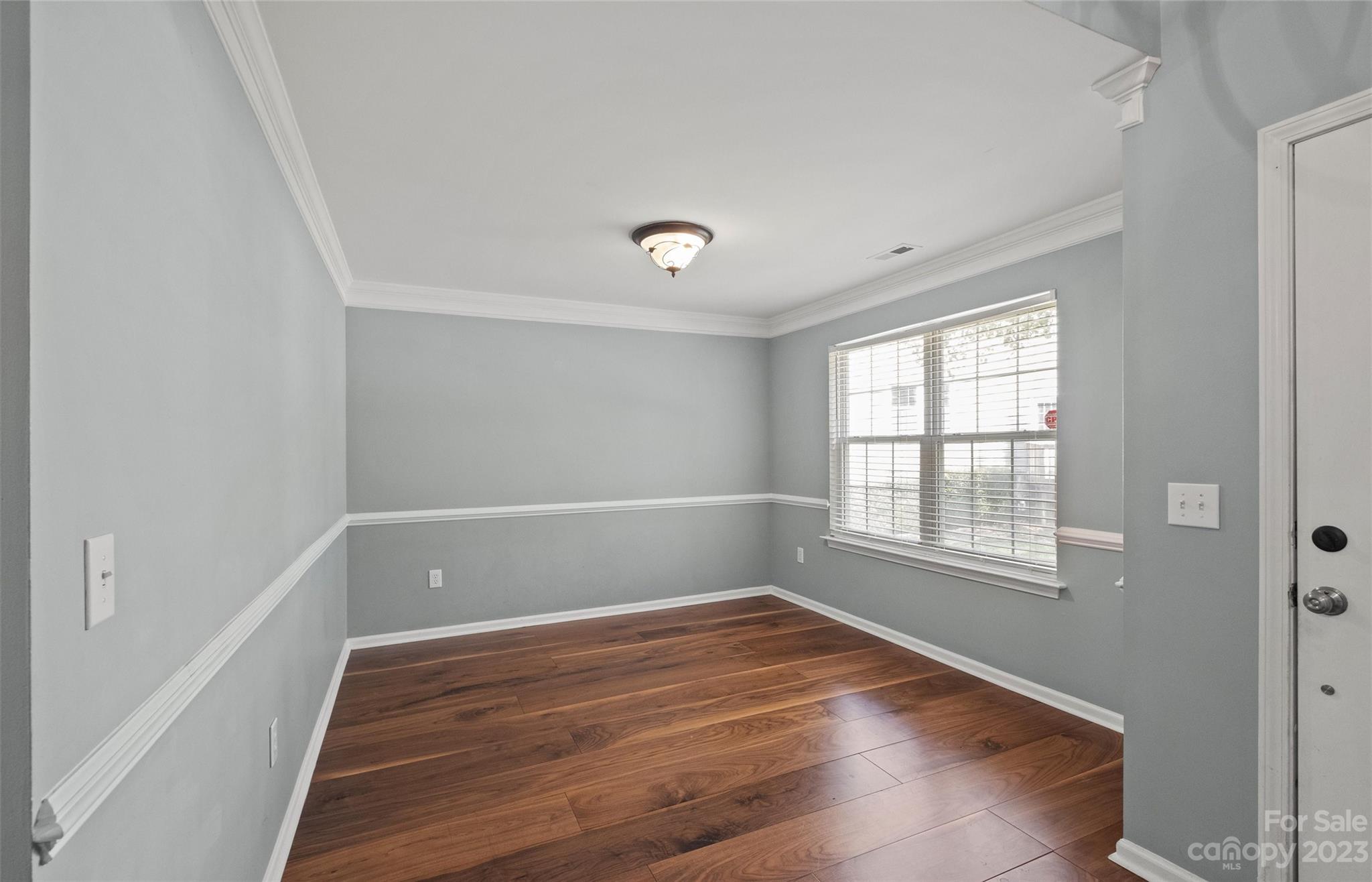 Empty gray room with dark wood floors, white molding, and a large window with blinds.