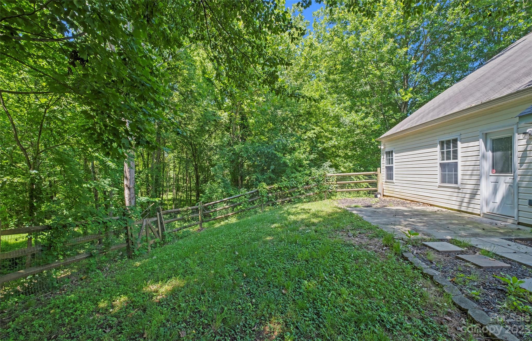 Wooded side yard beside a light-colored house, with a fenced lawn and concrete patio.