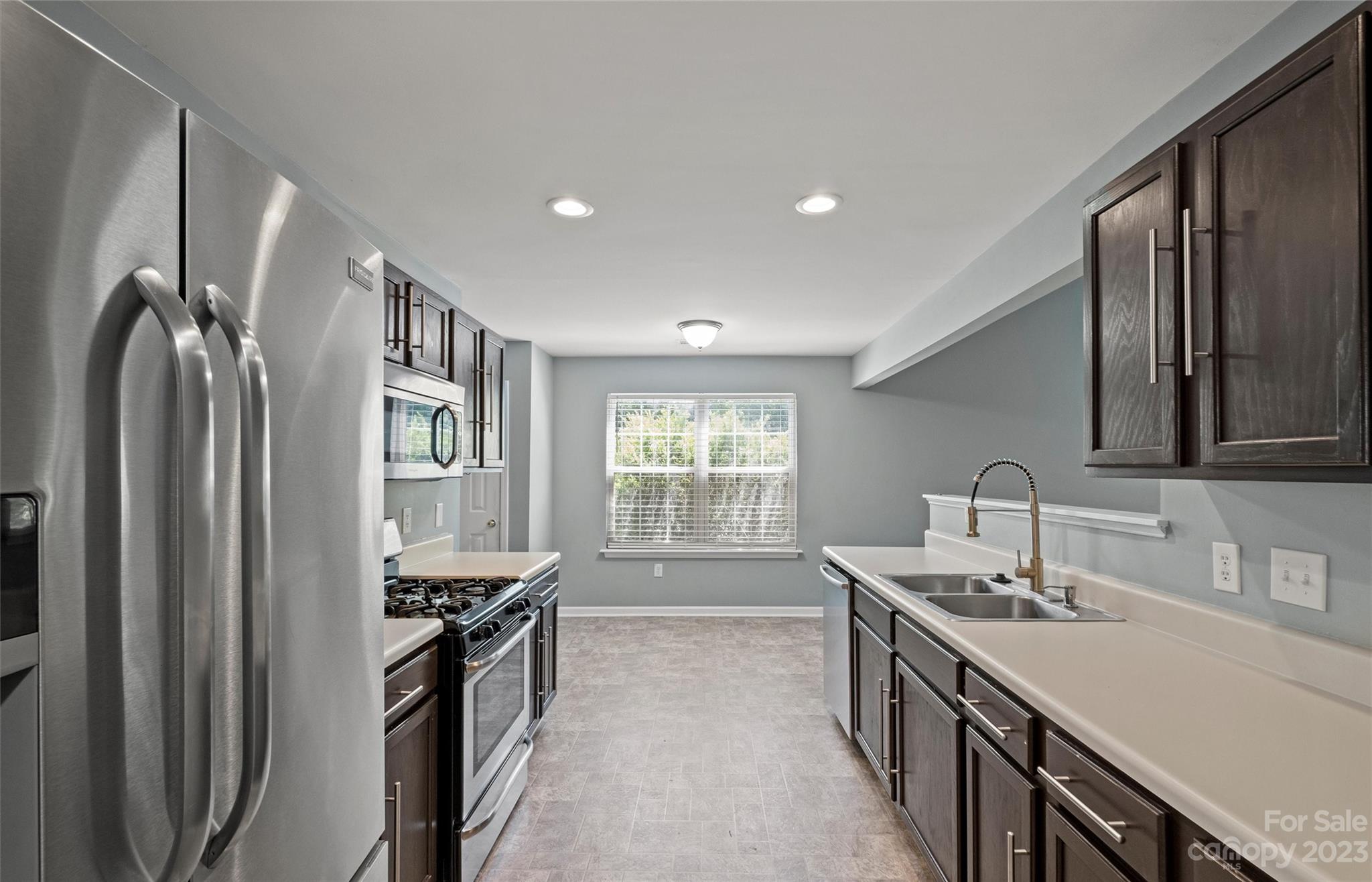 Galley kitchen with dark cabinets, stainless steel appliances, and a double sink beneath a window.