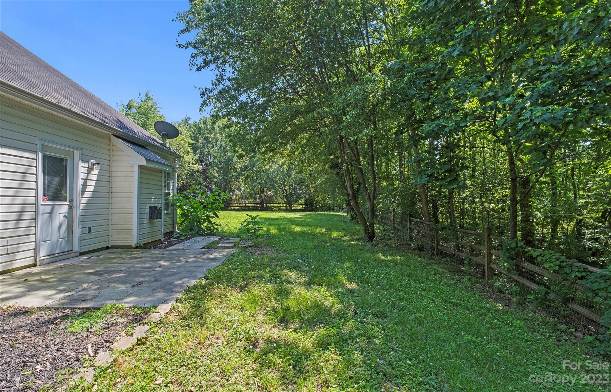 Backyard with a concrete patio, shaded lawn, mature trees, and a fence along the wooded edge.