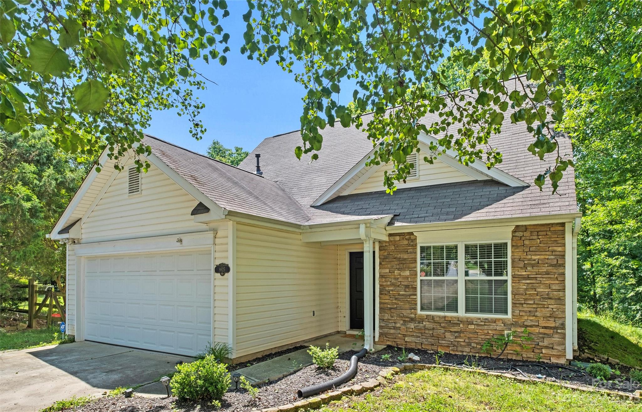Cream-sided home with a two-car garage, stone facade, and leafy trees overhead.