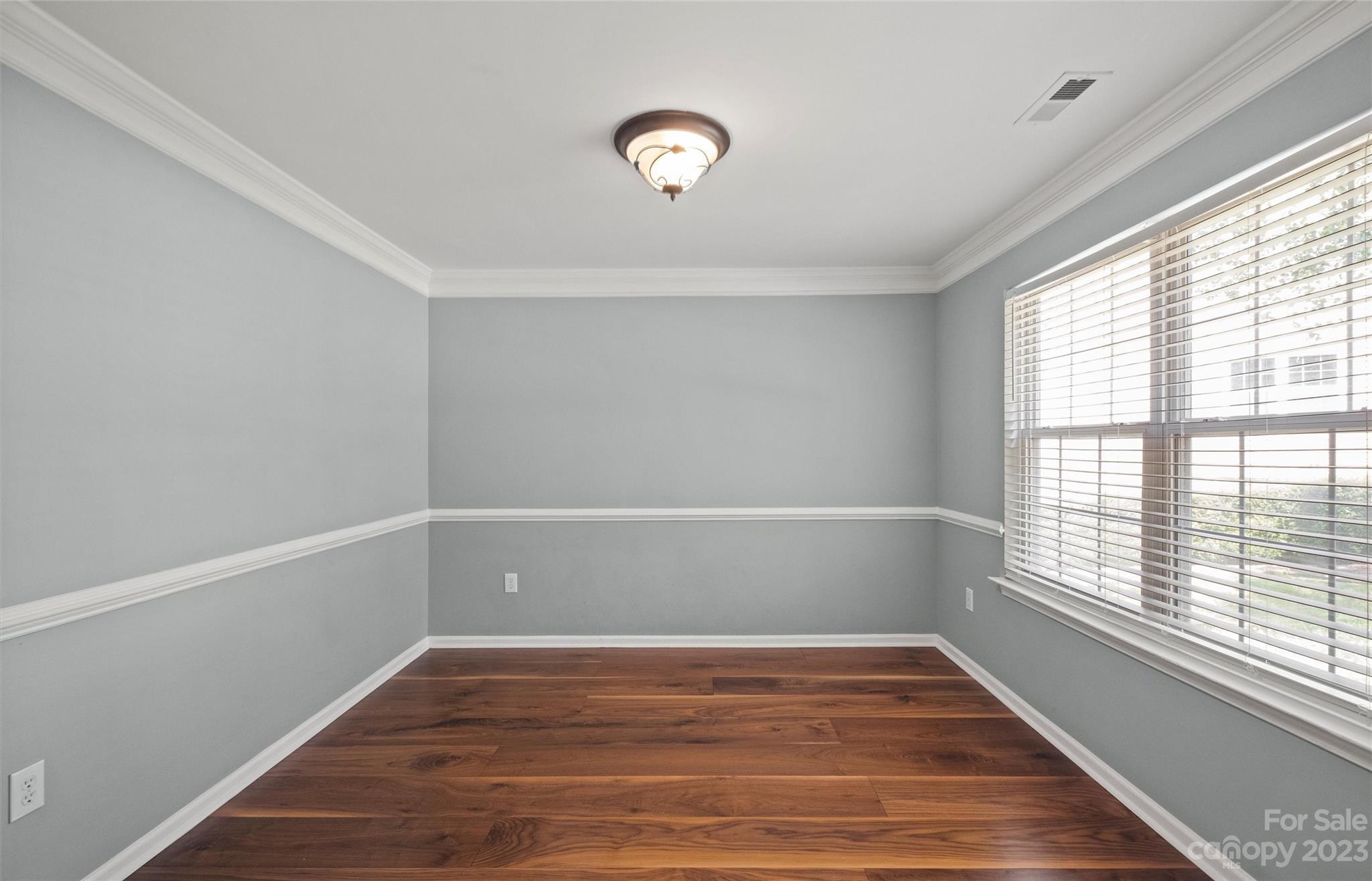 Empty gray room with dark hardwood floors, white trim, and a large window with blinds.