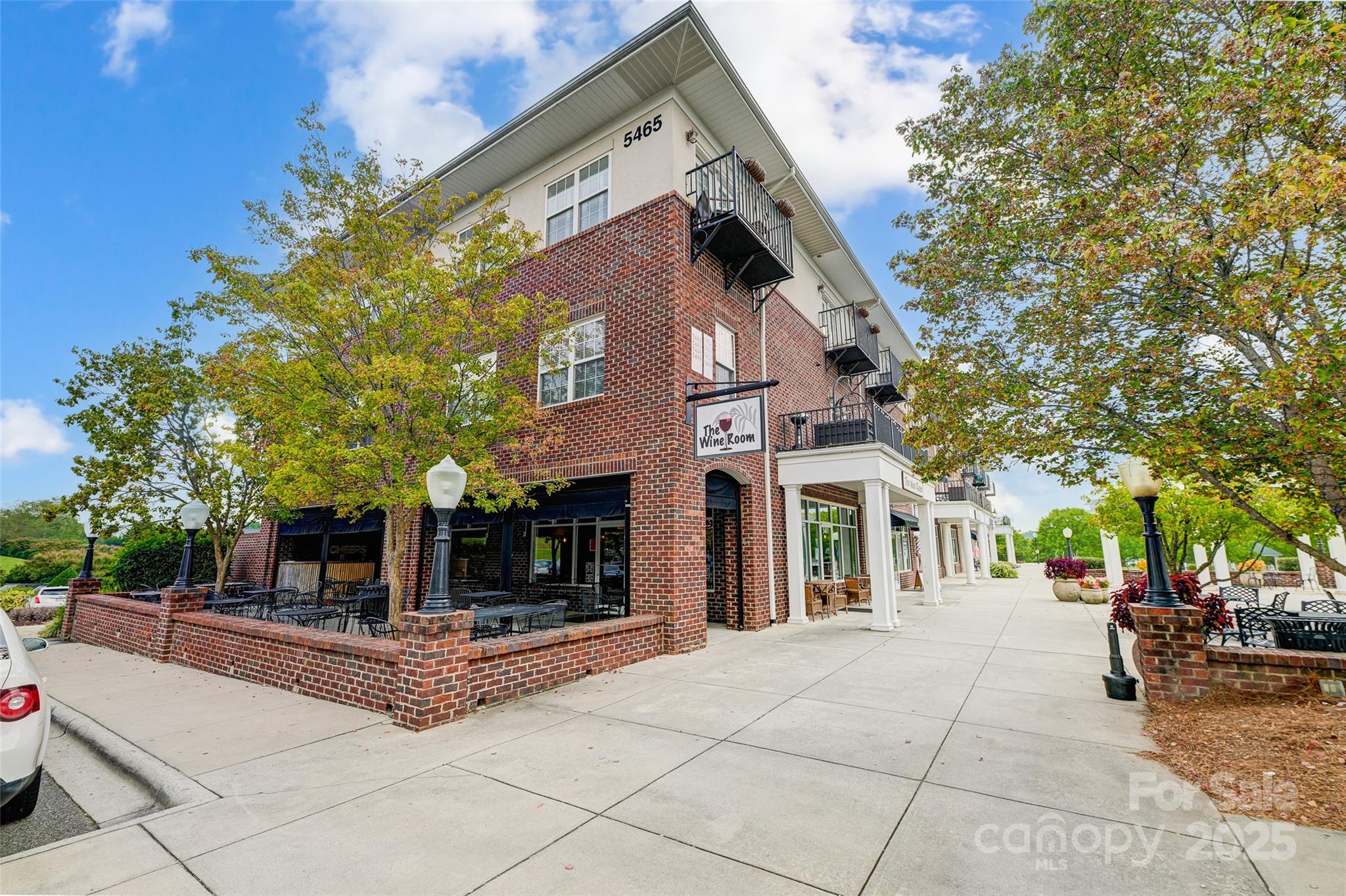 Brick commercial building with upper-floor balconies, restaurant patios, and a sidewalk beneath leafy trees.