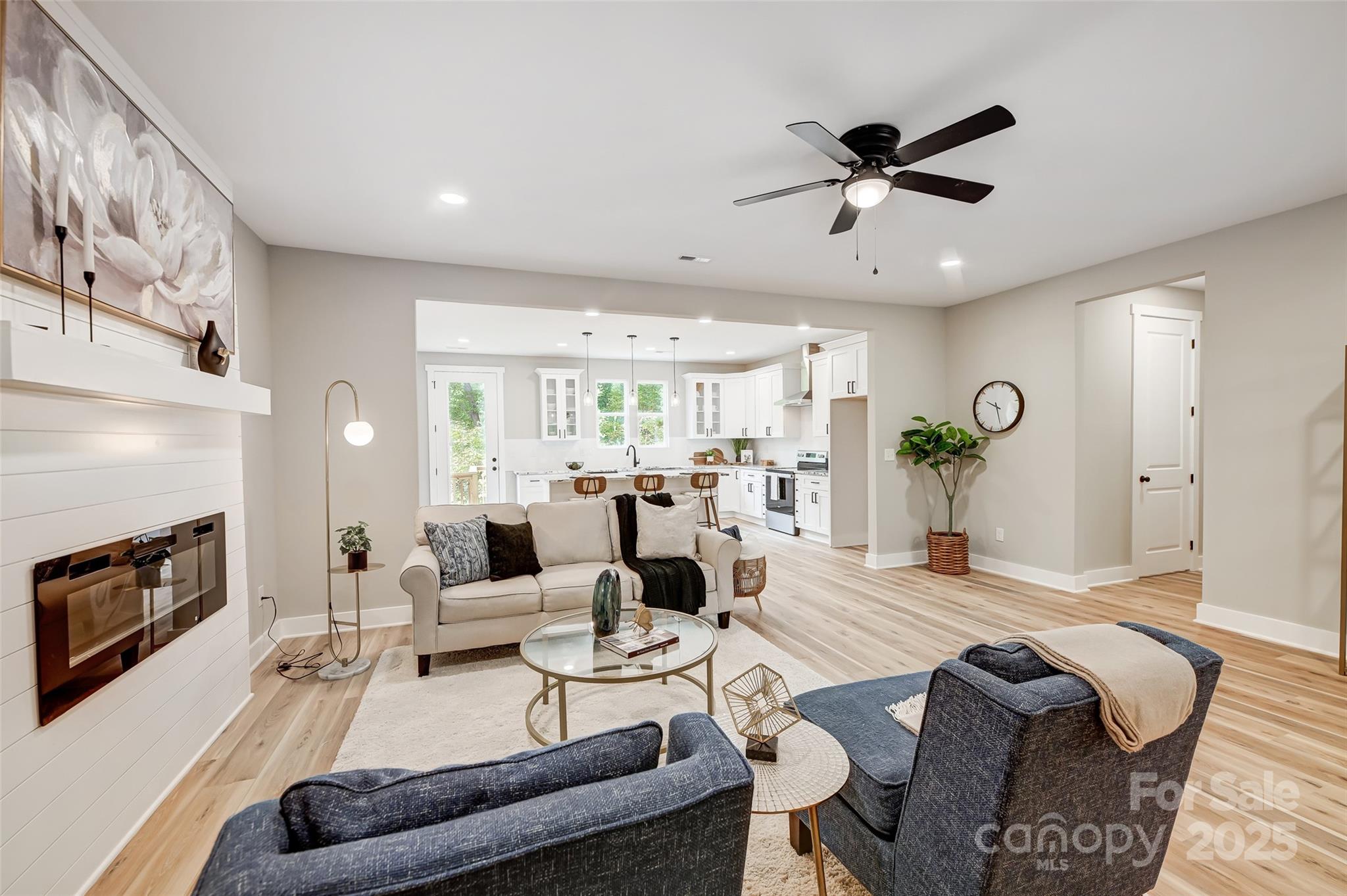 Staged living room opens to a bright white kitchen, with a fireplace and light wood floors.