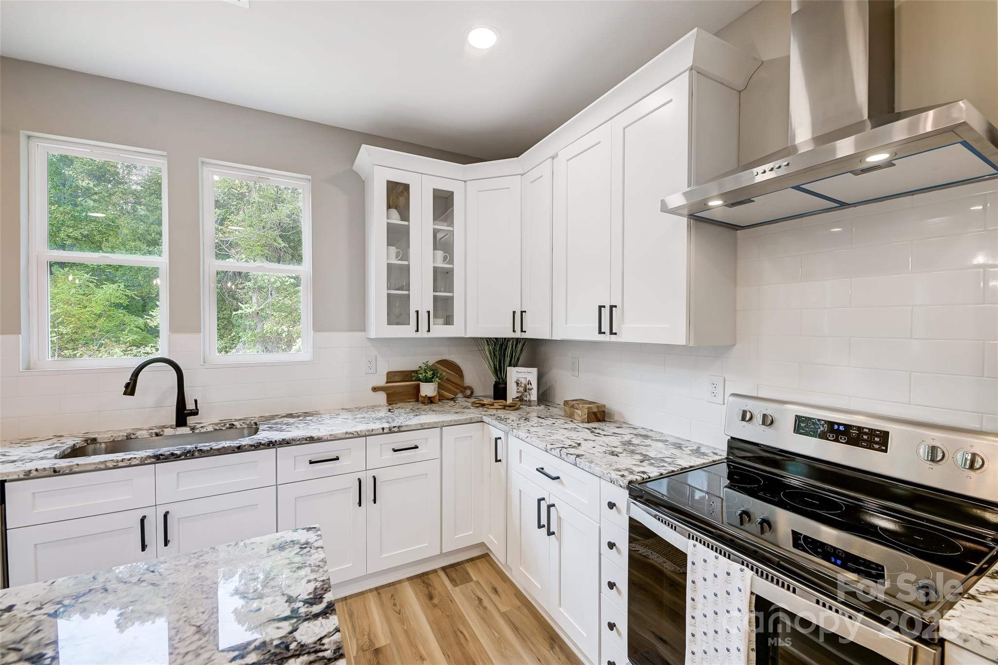 Kitchen with white cabinets, marble-patterned counters, stainless steel range, and two windows overlooking trees.