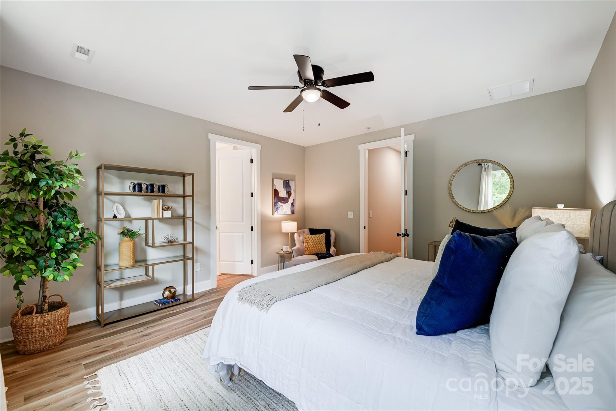 Staged bedroom with white bedding, navy pillows, wood-look floors, and a ceiling fan.