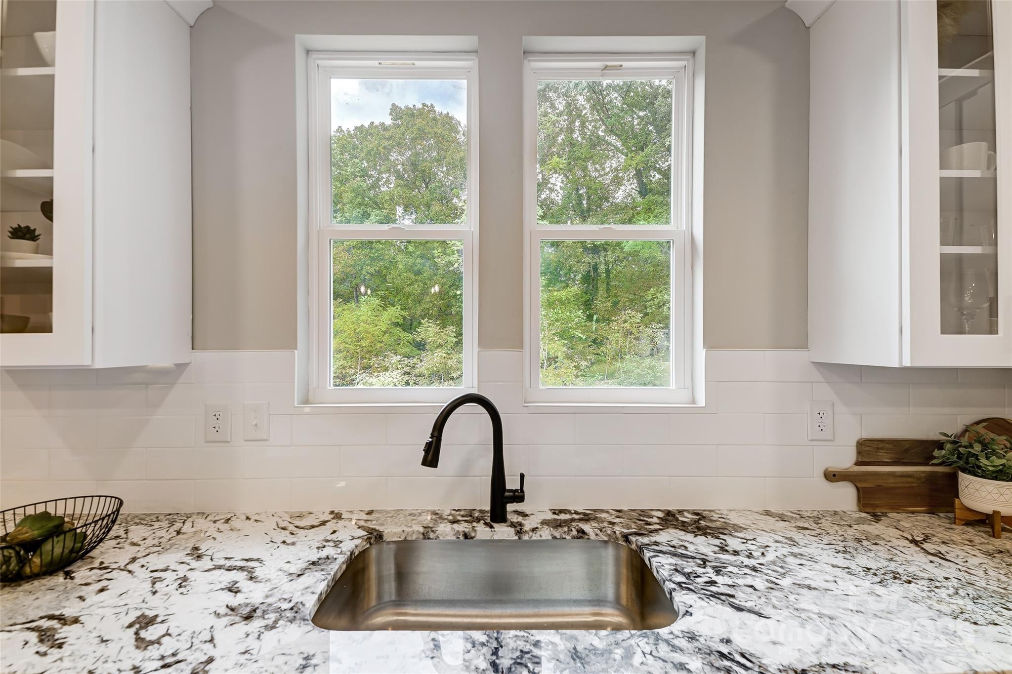 Kitchen sink and black faucet beneath two windows overlooking leafy trees.