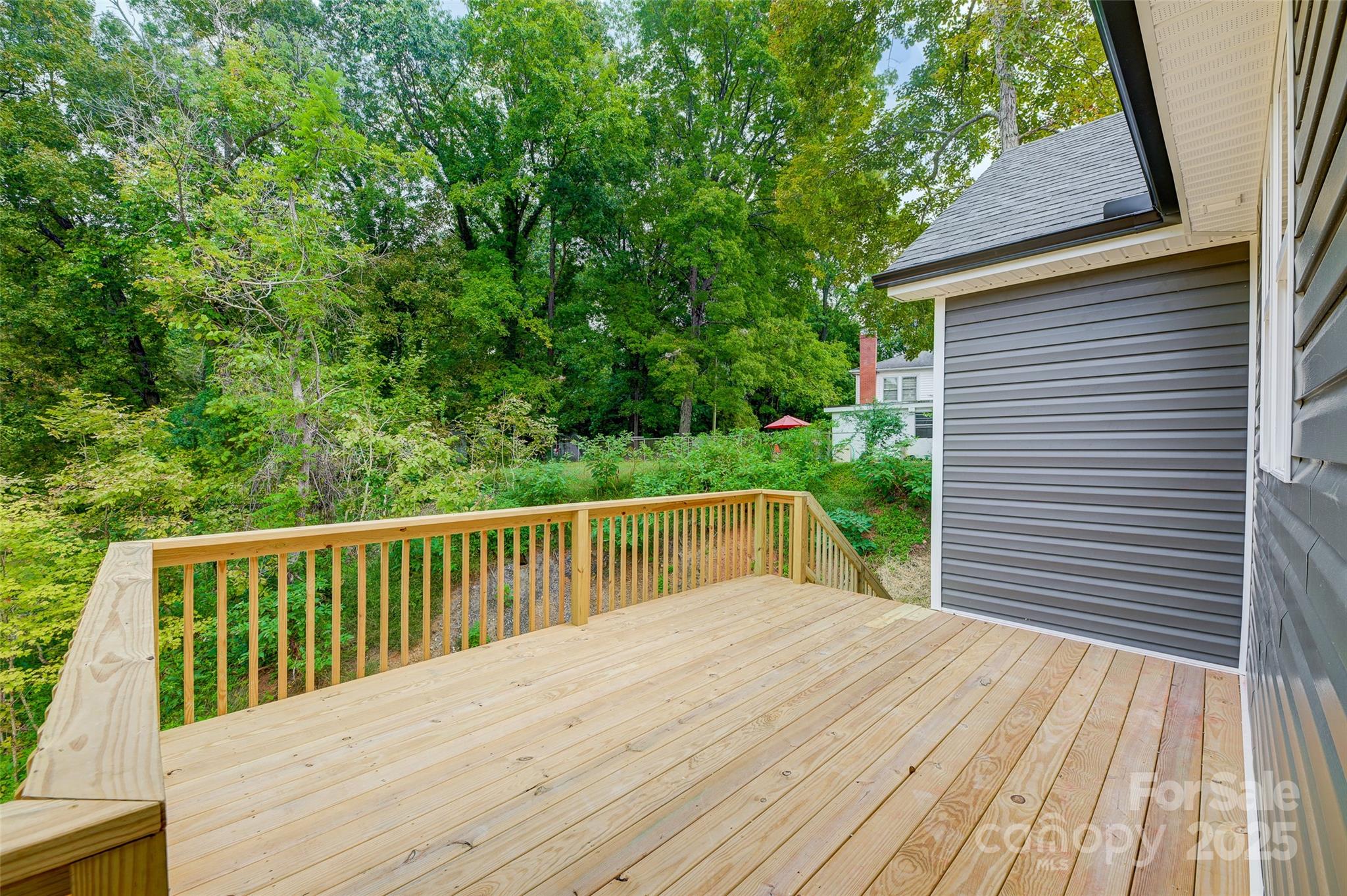 Wooden deck beside a gray-sided home, overlooking a densely wooded backyard.