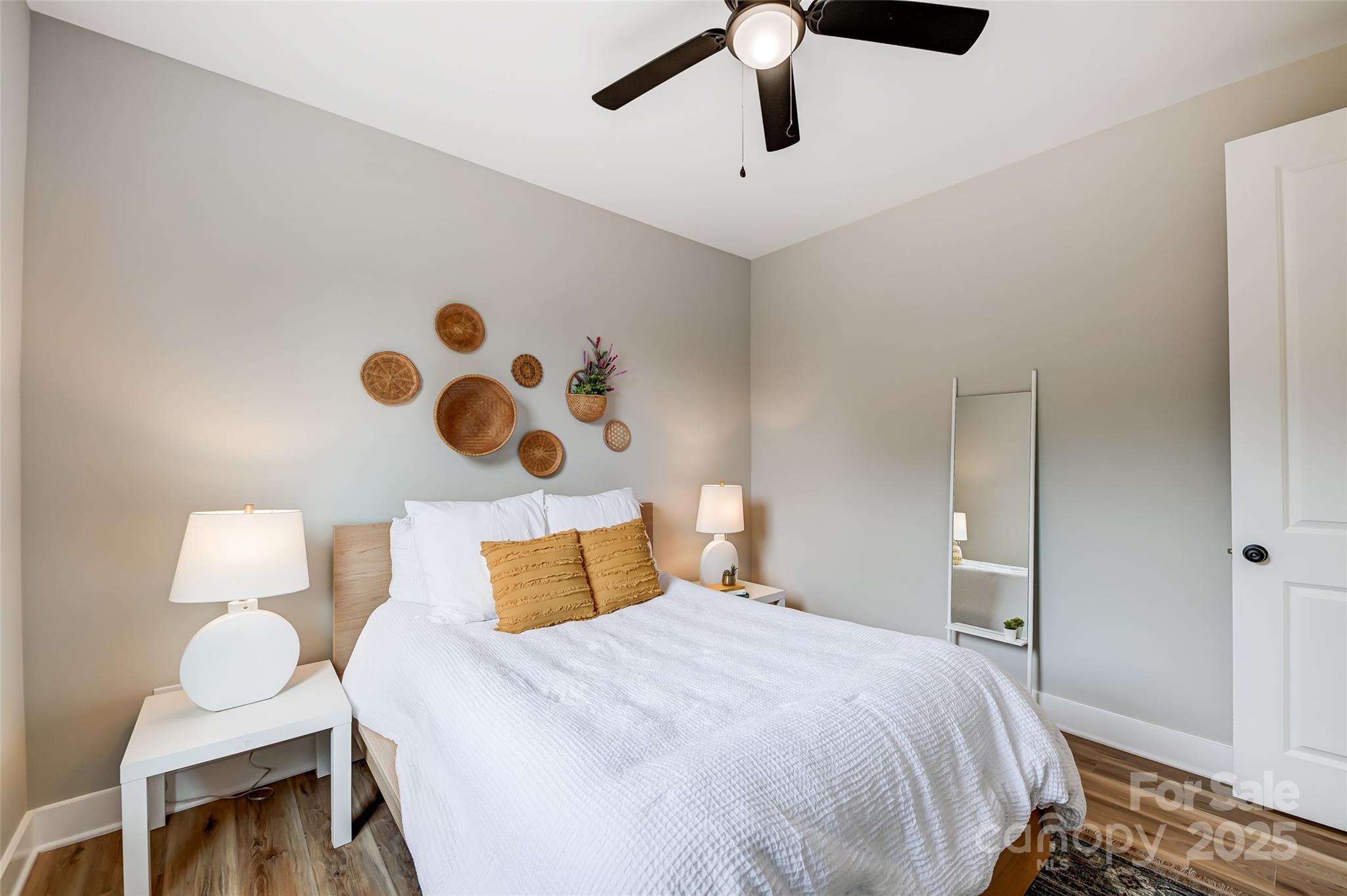 Staged bedroom with a white bed, mustard pillows, woven wall baskets, and a full-length mirror.