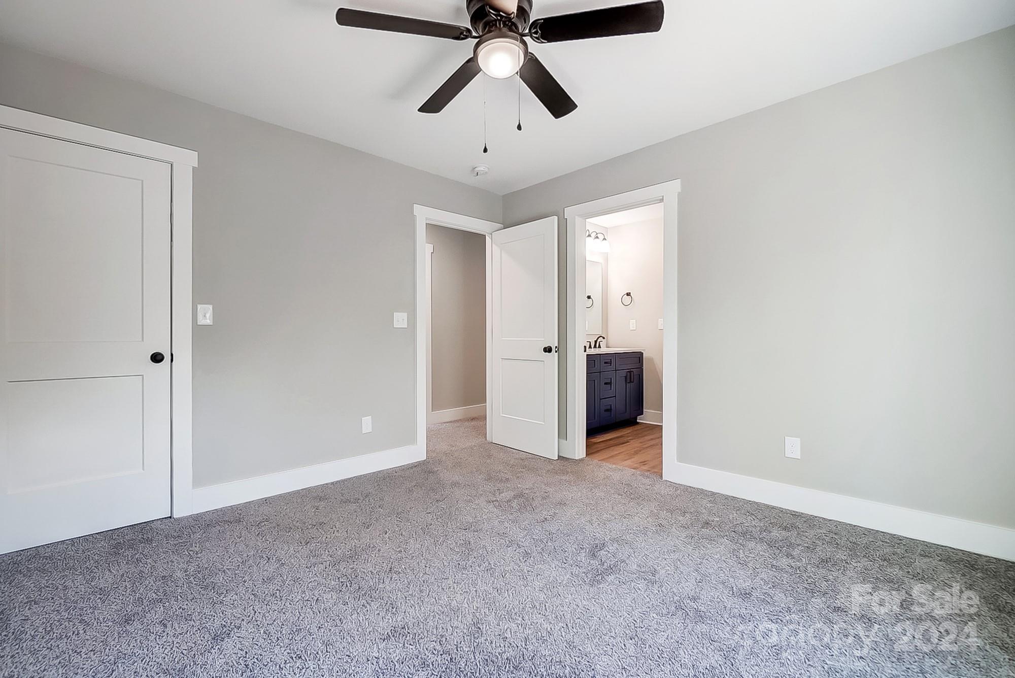 Empty carpeted bedroom with a ceiling fan and an open doorway to a bathroom with a dark vanity.
