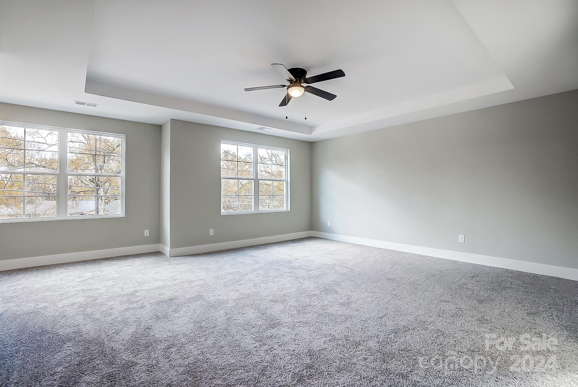 Empty carpeted bedroom with a tray ceiling, ceiling fan, and two sets of windows.