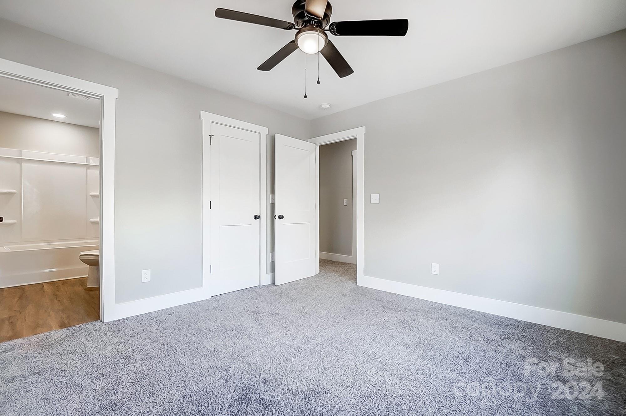 Vacant bedroom with gray carpet and ceiling fan, adjoining a bathroom and hallway.