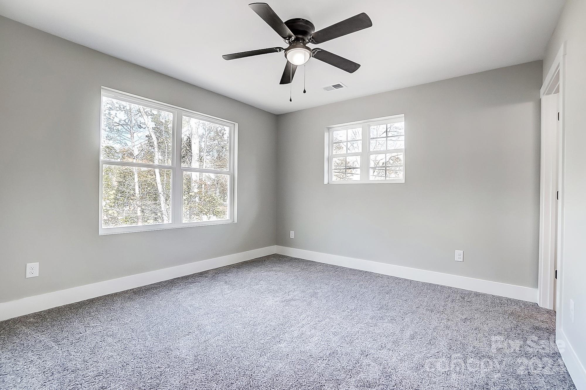 Empty bedroom with gray carpet, two windows, and a ceiling fan.