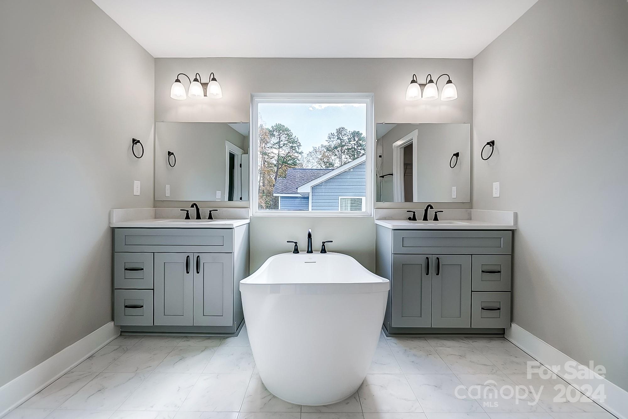 Bathroom with a freestanding soaking tub between matching gray double-sink vanities beneath a window.