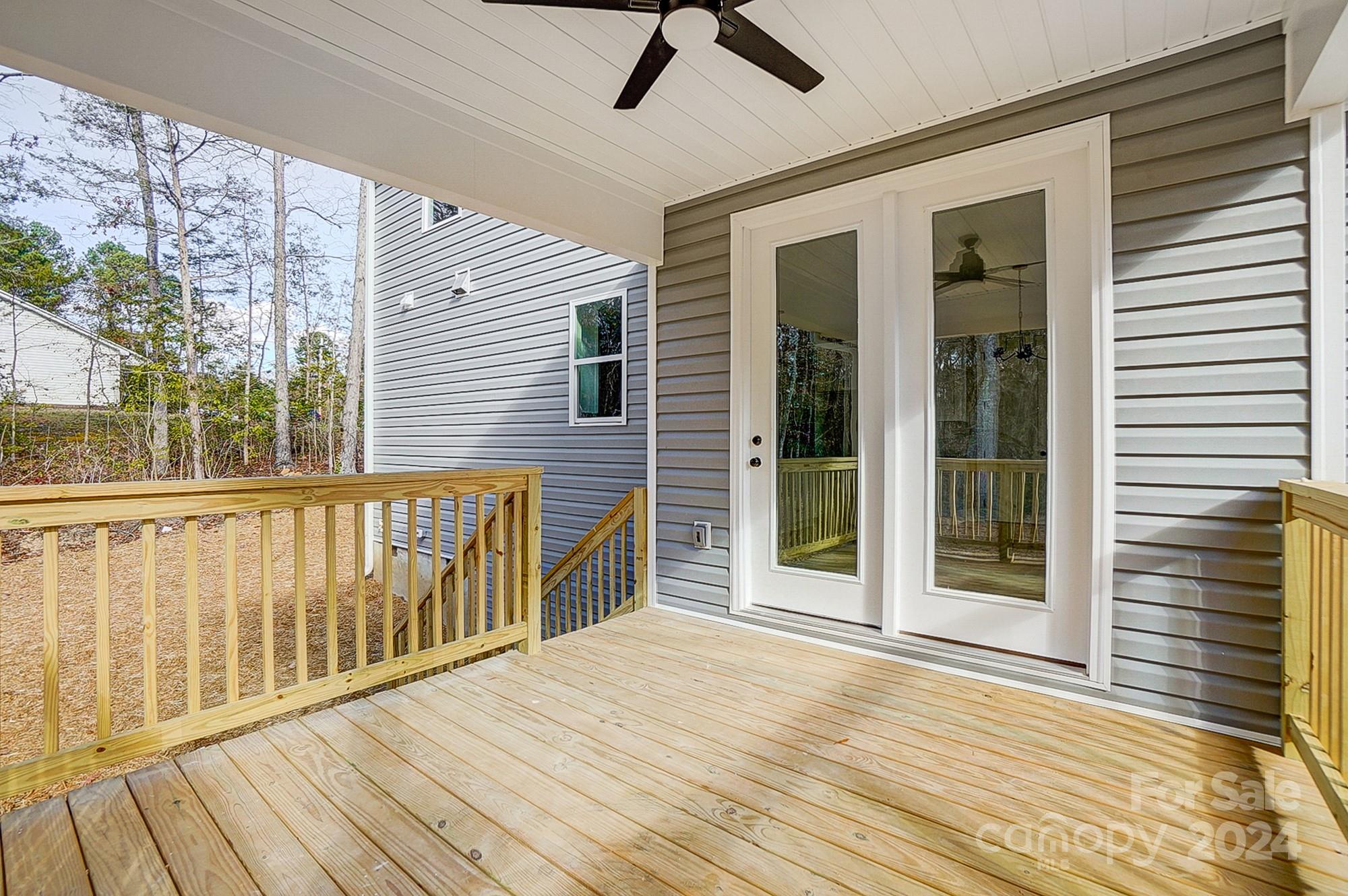 Covered wooden deck with railings, double glass doors, and a ceiling fan at the back of a gray-sided home.