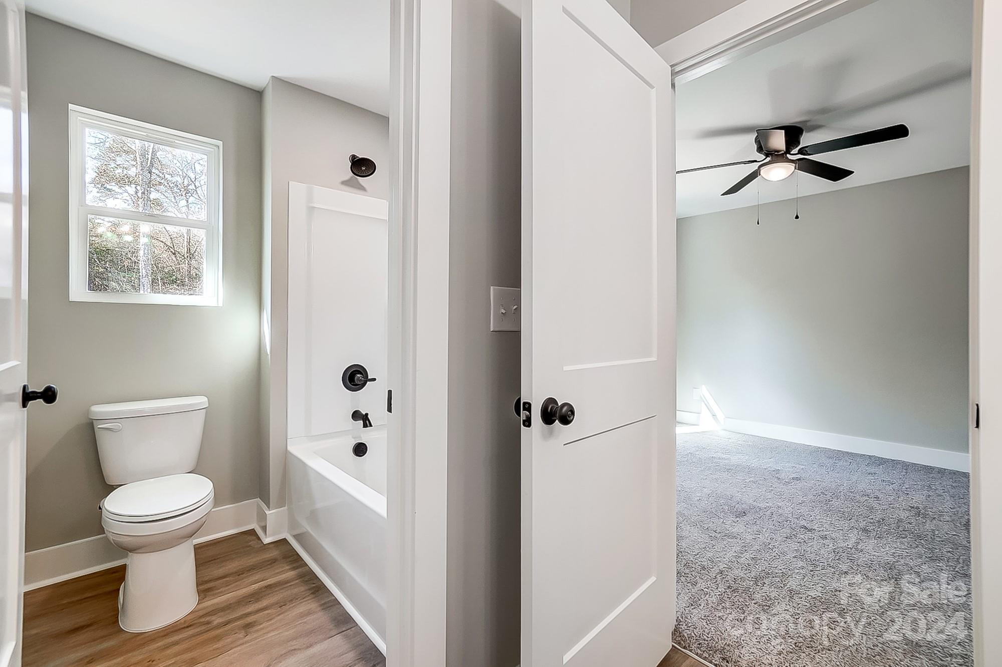 Bathroom with a window above the toilet opens into an empty carpeted bedroom with a ceiling fan.