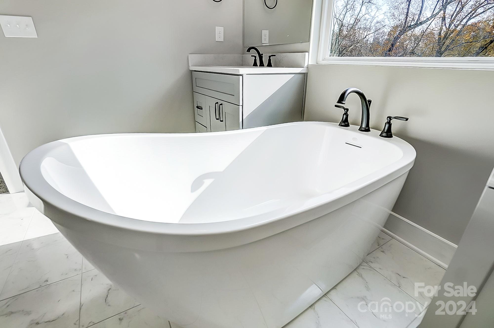 Bathroom with a large freestanding soaking tub, black faucets, and a window overlooking autumn trees.