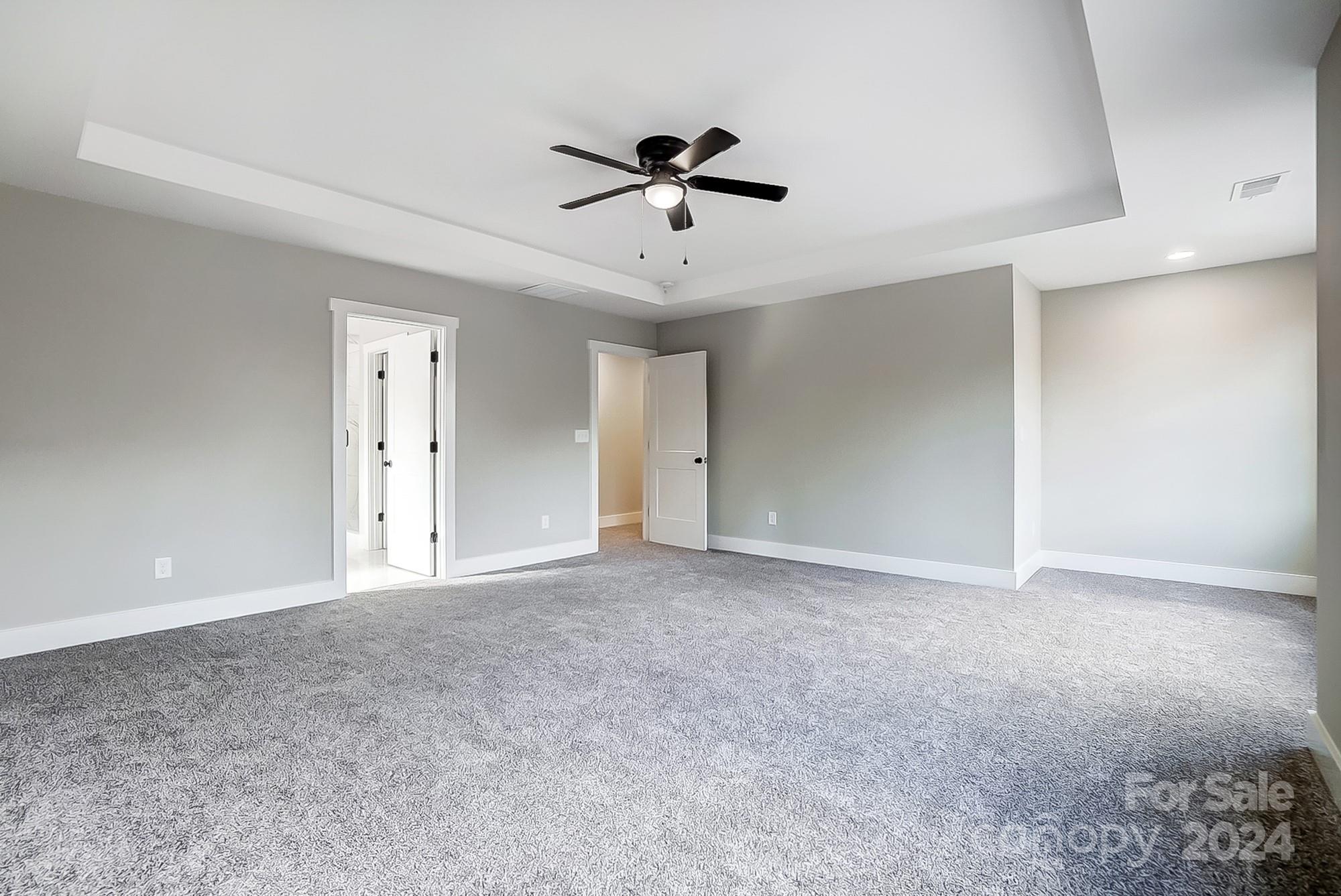 Vacant bedroom with gray carpet, a tray ceiling, and a dark ceiling fan with a light.