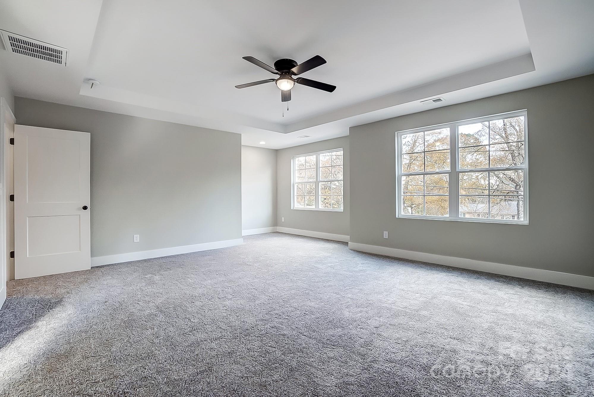 Empty bedroom with gray carpet, tray ceiling, black ceiling fan, and two large windows.
