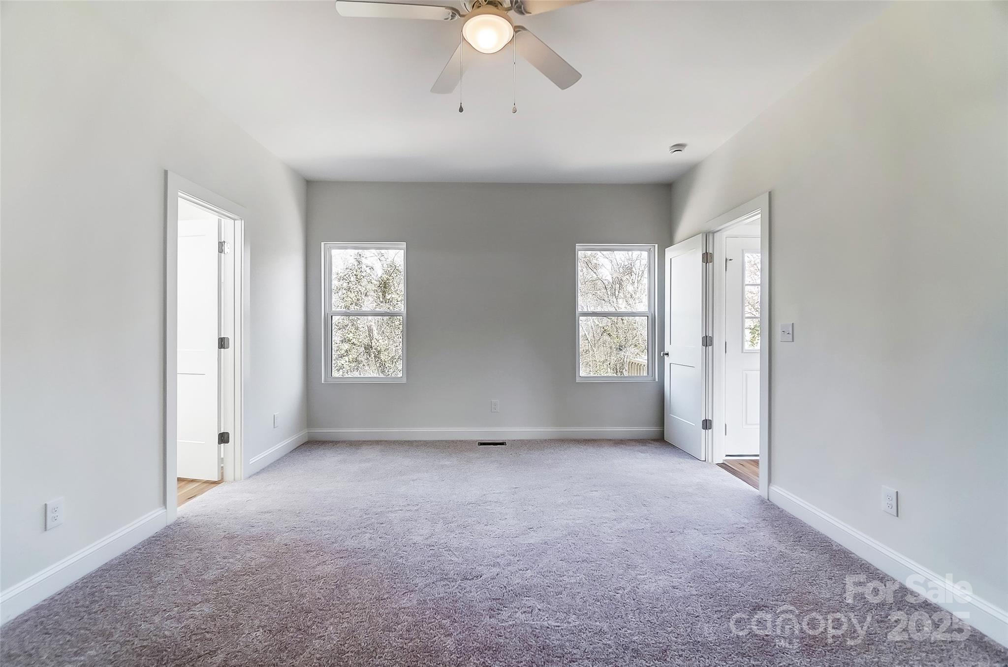 Empty bedroom with gray carpeting, a ceiling fan, and two windows on the far wall.
