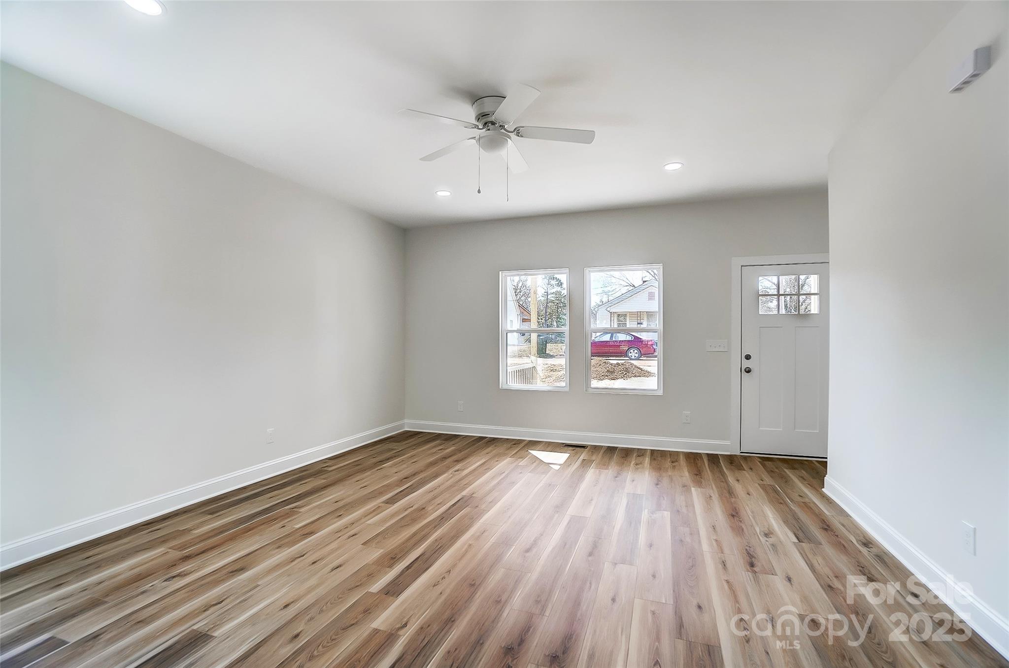 Empty front room with wood-look plank flooring, ceiling fan, two windows, and an entry door.