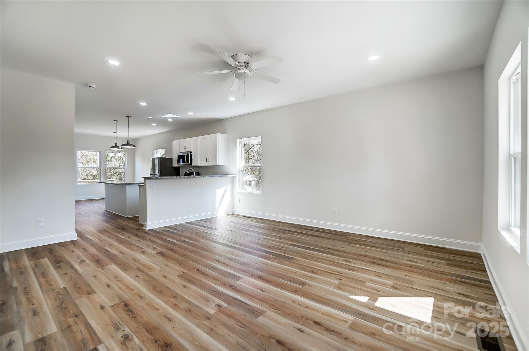 Vacant open-plan living area with wood-look floors, ceiling fan, and adjoining white kitchen.