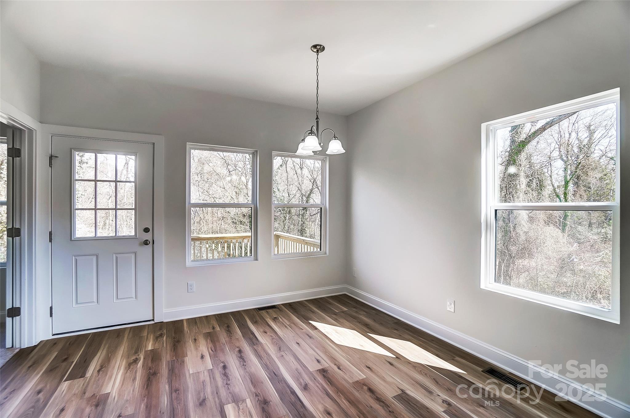 Empty dining area with wood-look flooring, a glass-paneled door, and windows overlooking leafless trees.