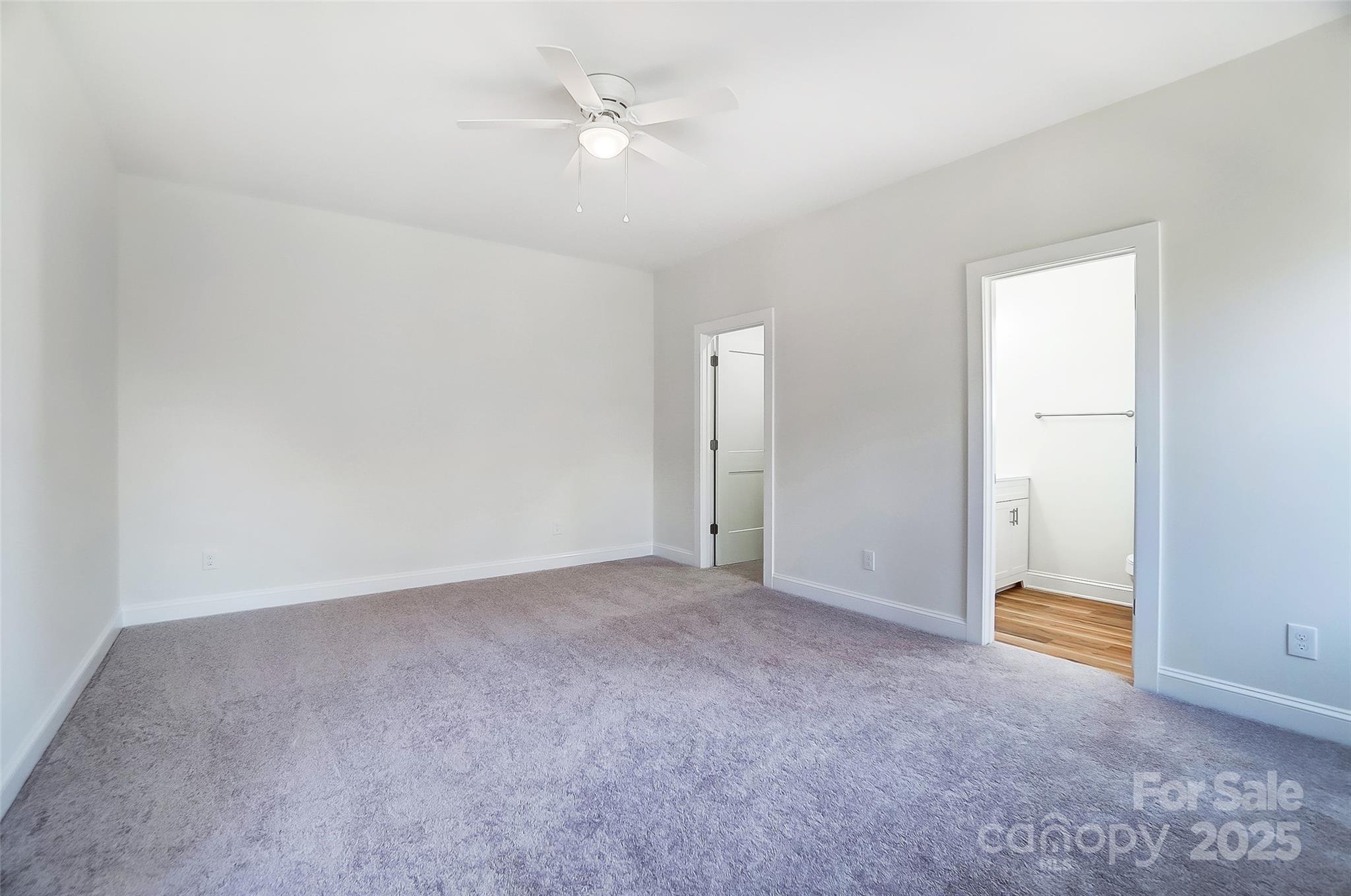 Empty bedroom with gray carpet and ceiling fan, opening to a bathroom with wood-look flooring.