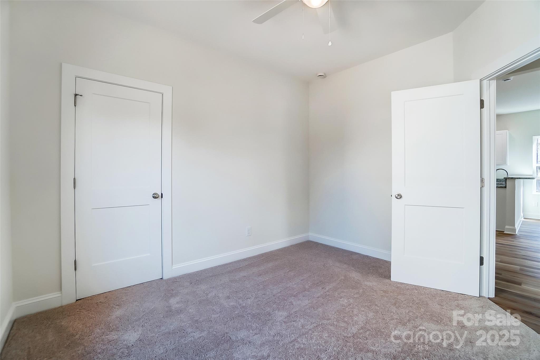 Empty carpeted bedroom with white walls, a ceiling fan, and an open doorway to a wood-floored room.