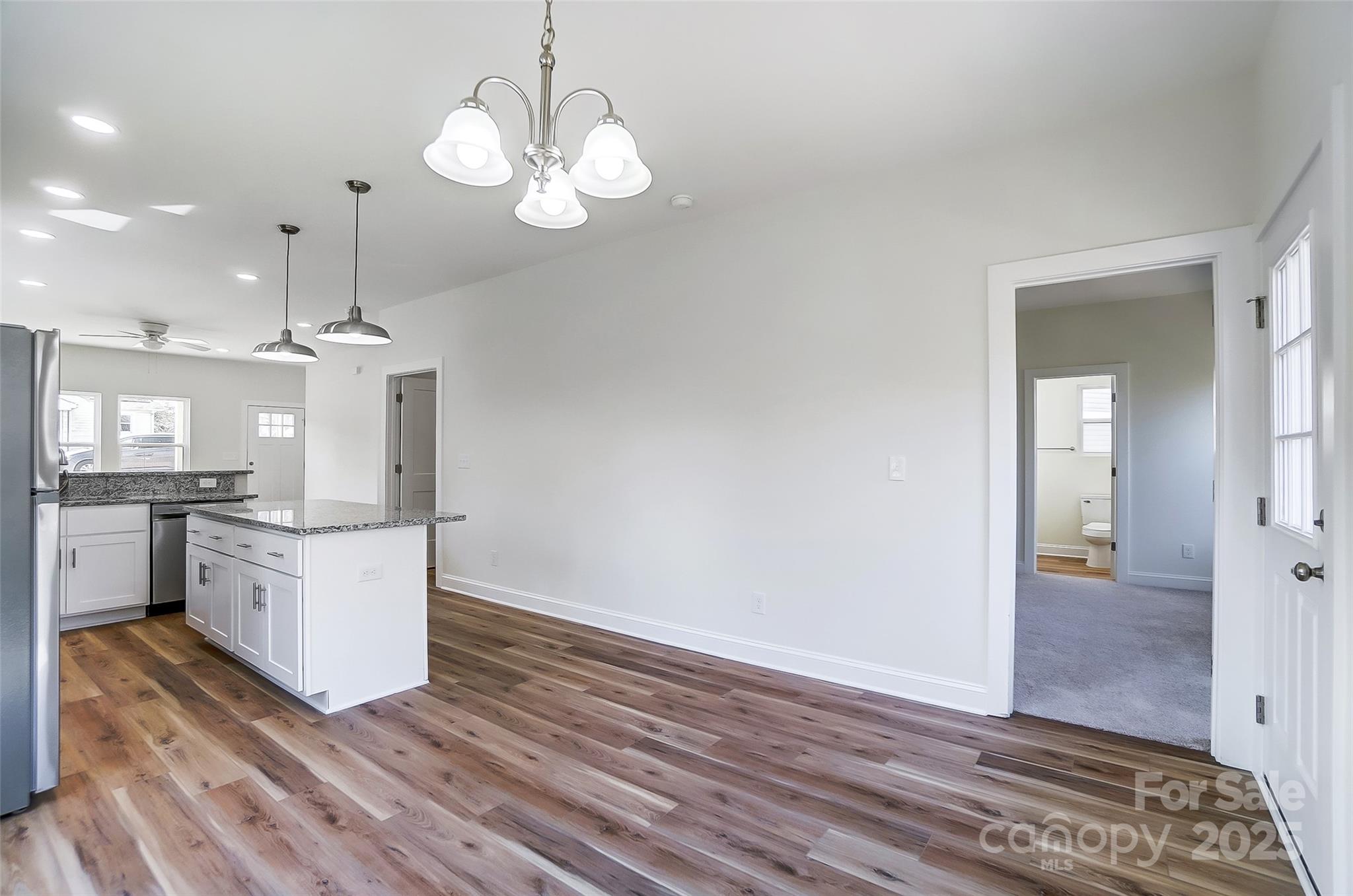 Open kitchen and dining area with granite island, wood-look flooring, and an adjoining carpeted room.