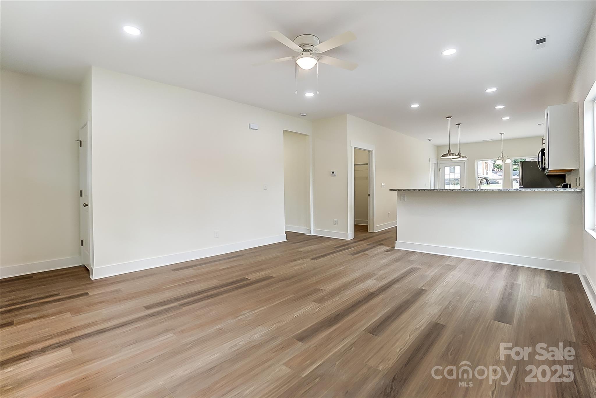 Empty open-plan living area with brown flooring, ceiling fan, and kitchen breakfast bar.