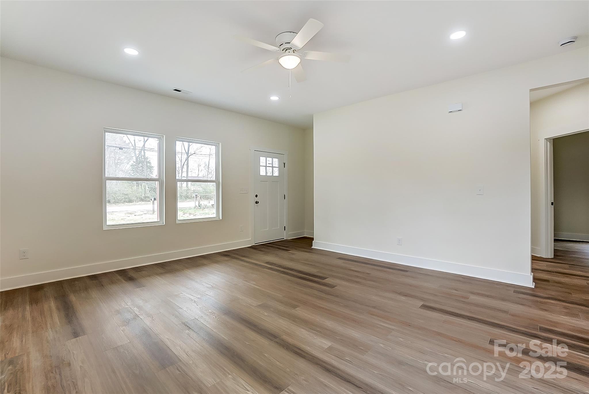 Vacant room with brown wood-look floors, two windows, a front door, and a ceiling fan.