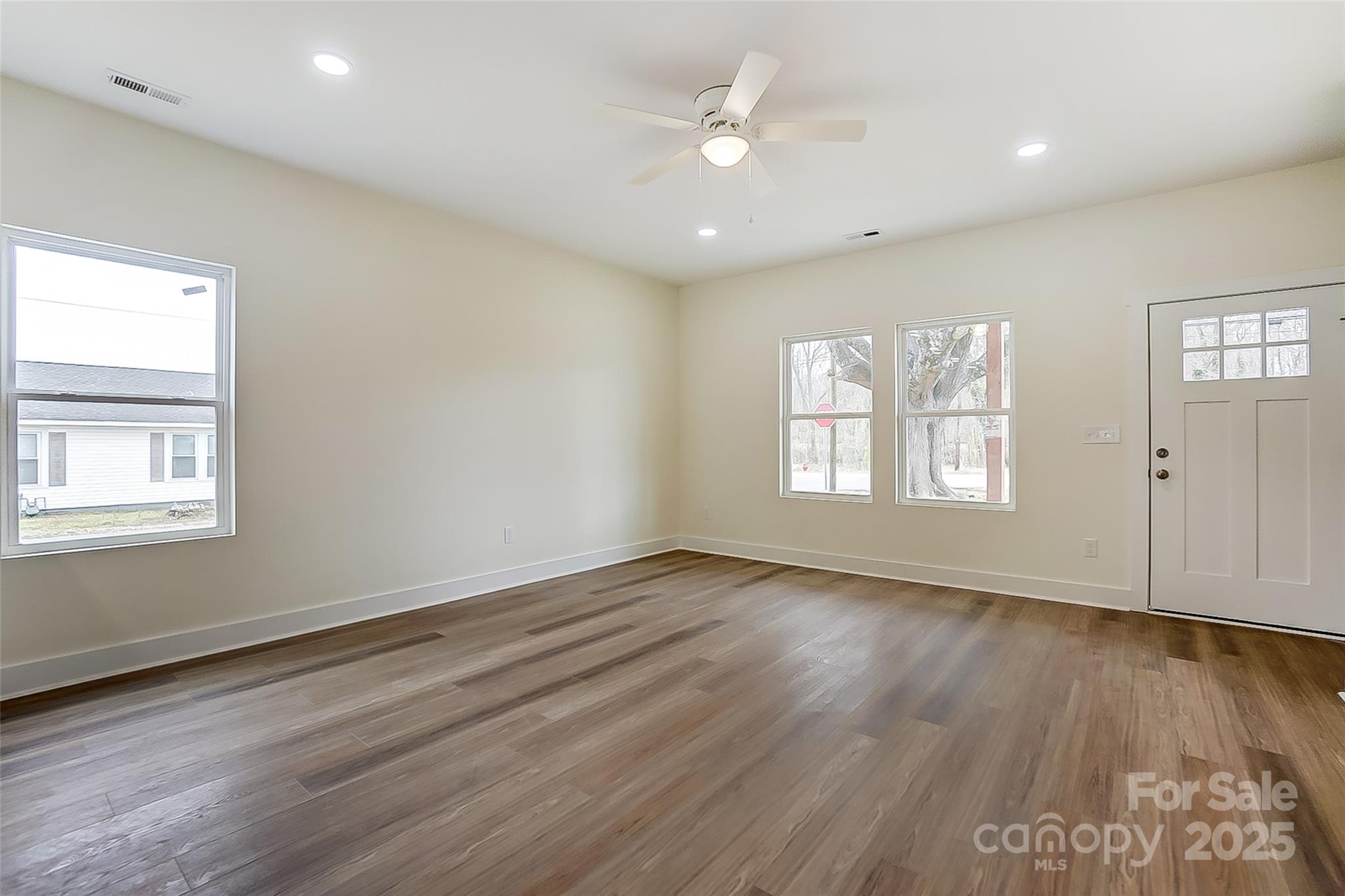 Vacant living room with wood-look floors, three windows, and a white front door.