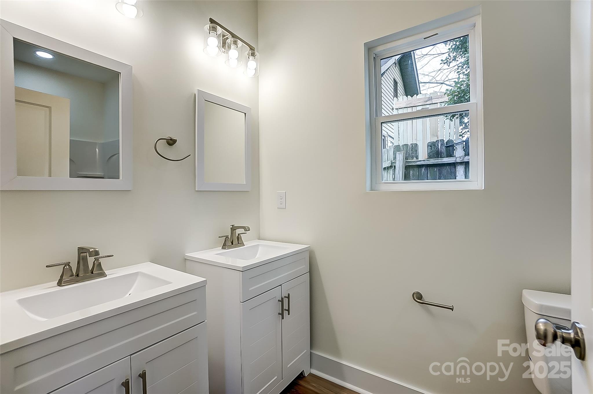 Bathroom with two white vanities, framed mirrors, and a window overlooking a fence.