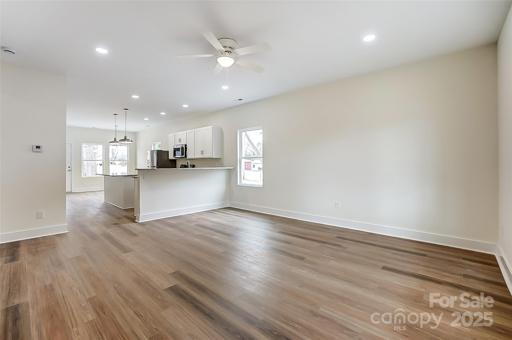 Empty open-plan living area with wood-look floors, white kitchen peninsula, and ceiling fan.