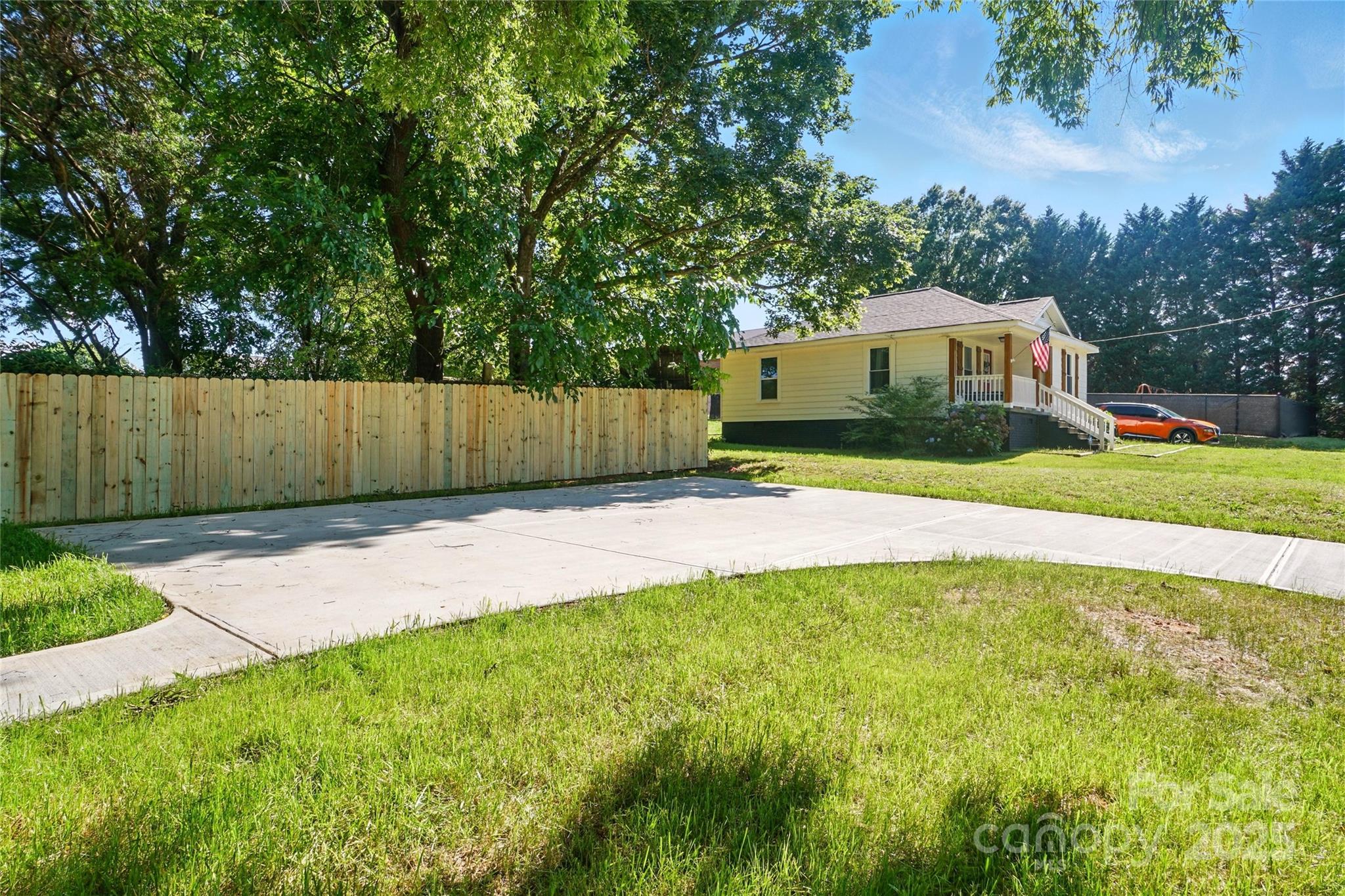Yellow single-story house with a covered porch, American flag, broad driveway, and wooden fence.