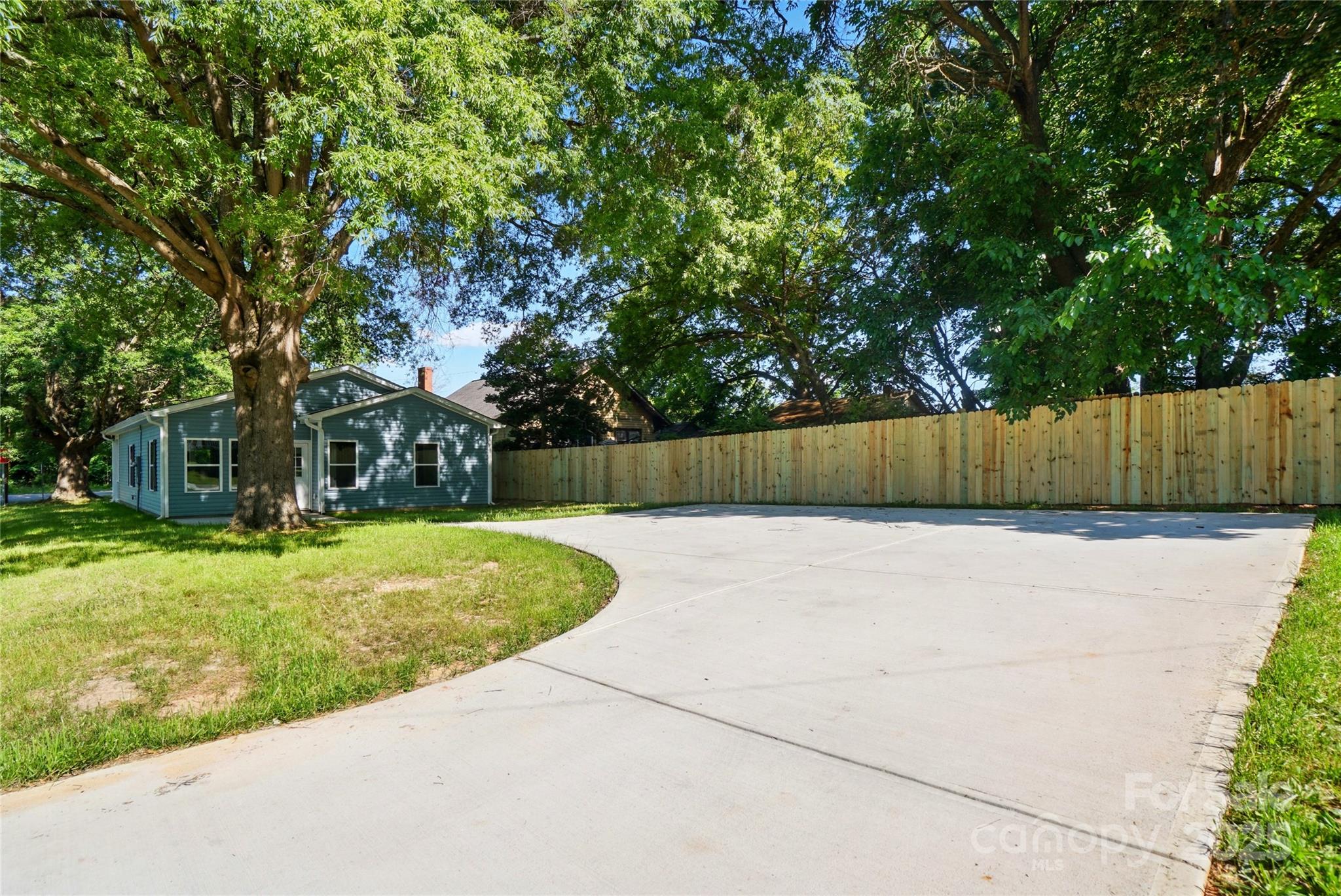 Blue-gray house beside a broad driveway and tall wooden fence beneath mature trees.