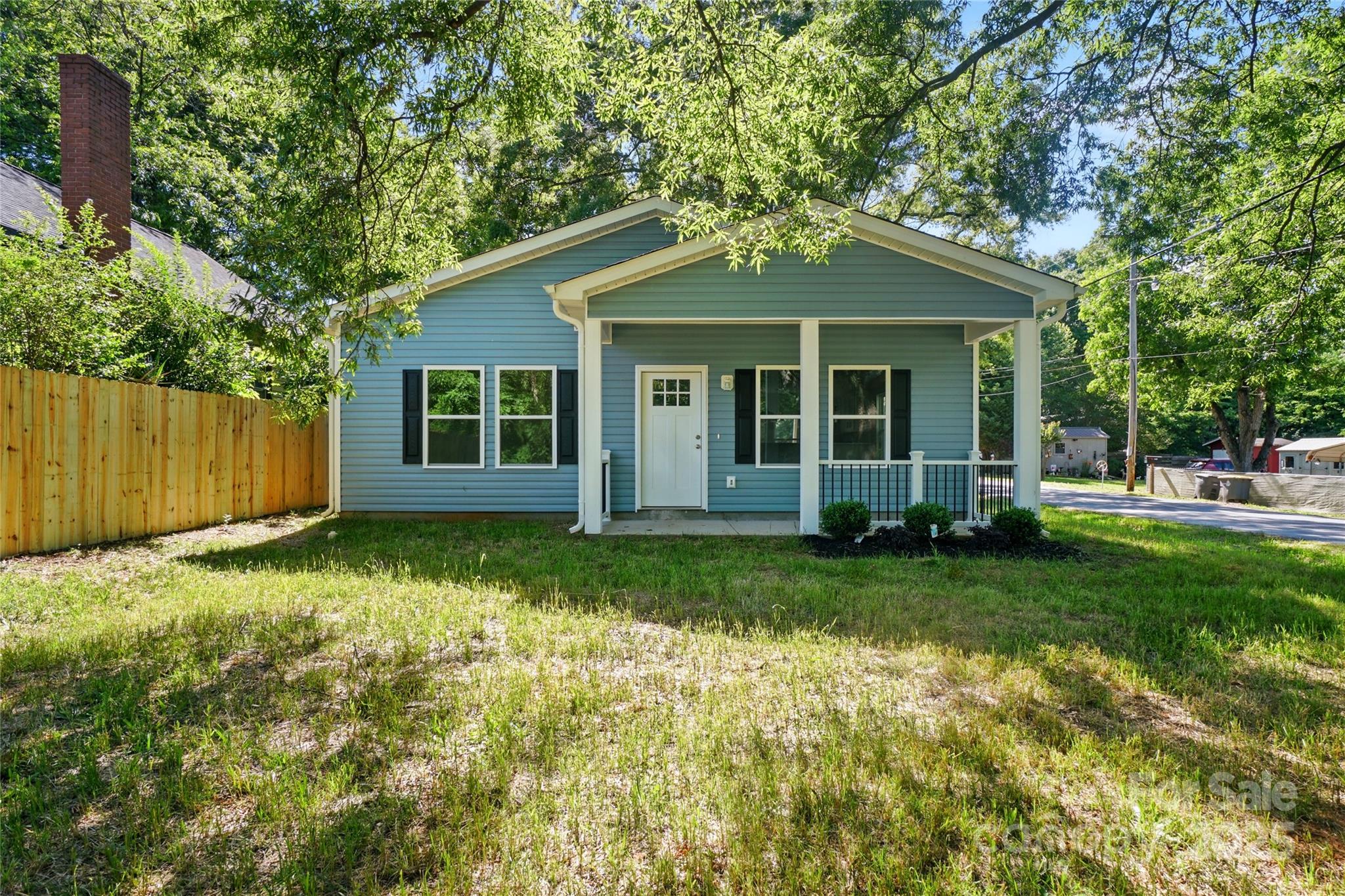 Blue single-story house with a covered porch and wooden fence beneath mature trees.