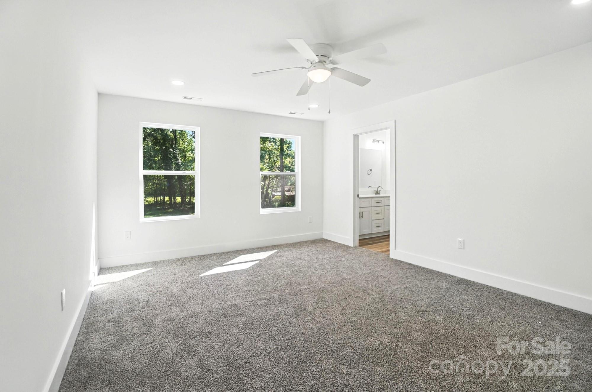 Empty carpeted bedroom with two tree-facing windows, a ceiling fan, and an open bathroom doorway.