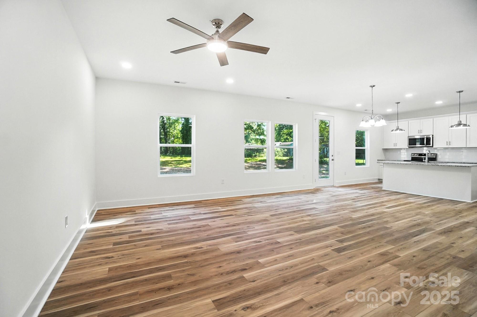 Vacant open living and dining area with wood floors, a ceiling fan, and an adjoining white kitchen.
