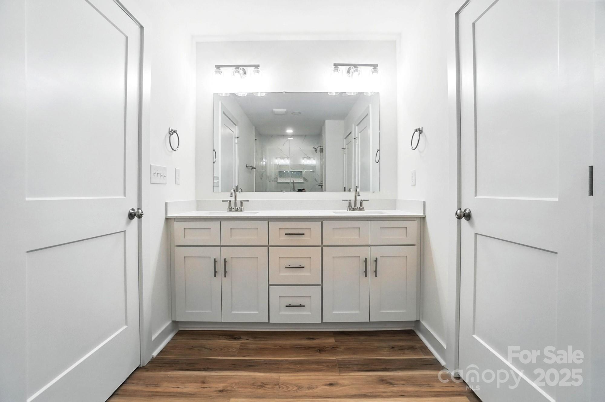 Bathroom with a white double-sink vanity, large mirror, and dark wood-look flooring.