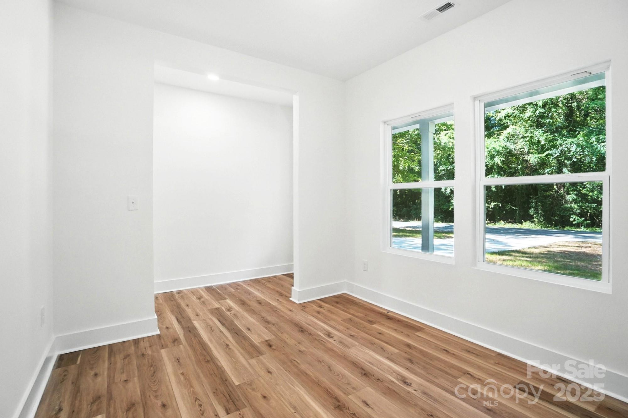 Empty room with warm wood floors and two large windows overlooking leafy trees.