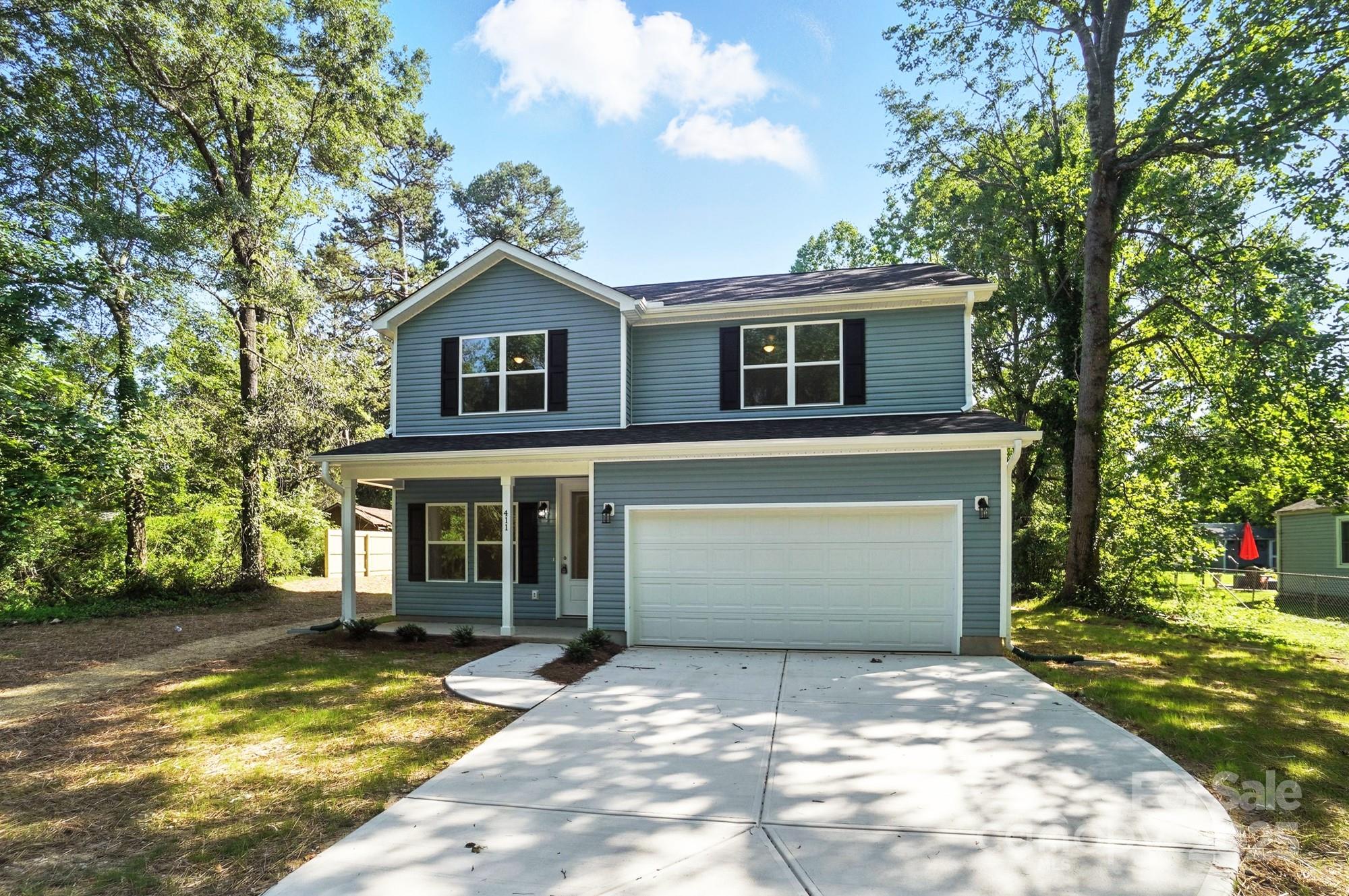 Two-story blue-gray house with a covered porch, double garage, and wide driveway among mature trees.