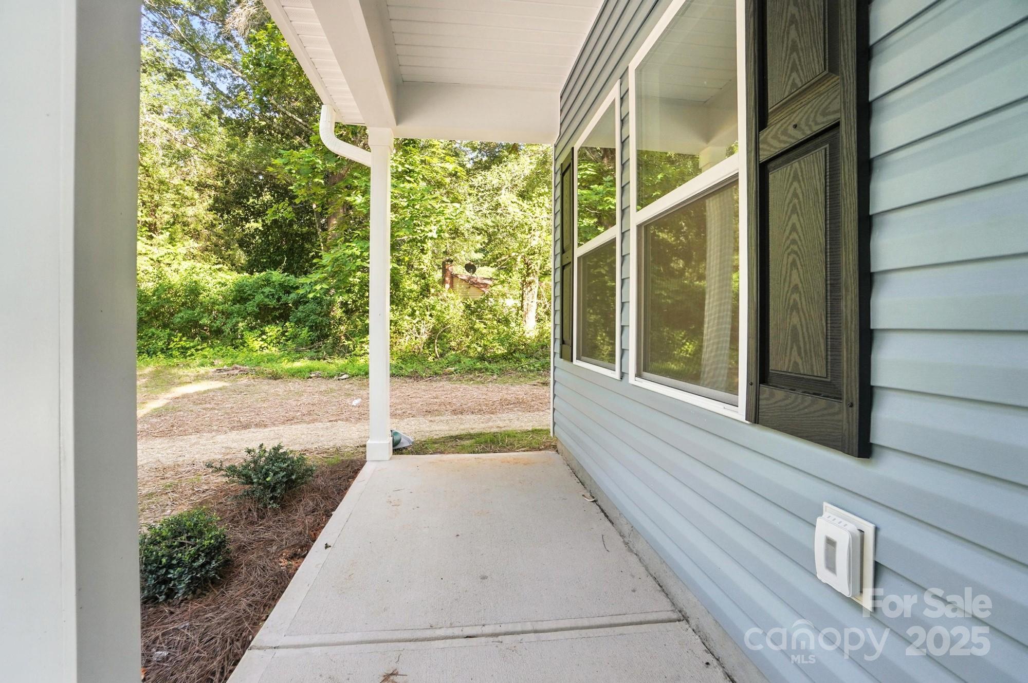 Covered concrete porch beside a light blue house, overlooking a leafy wooded yard.
