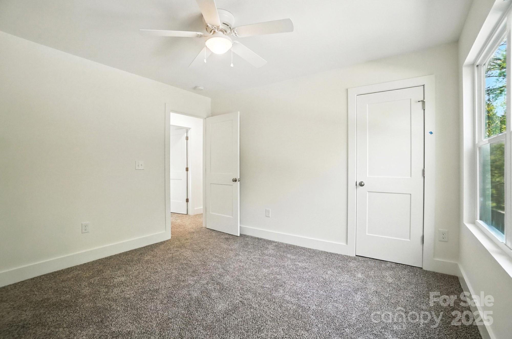 Empty bedroom with gray carpet, a ceiling fan, two white doors, and a large window.