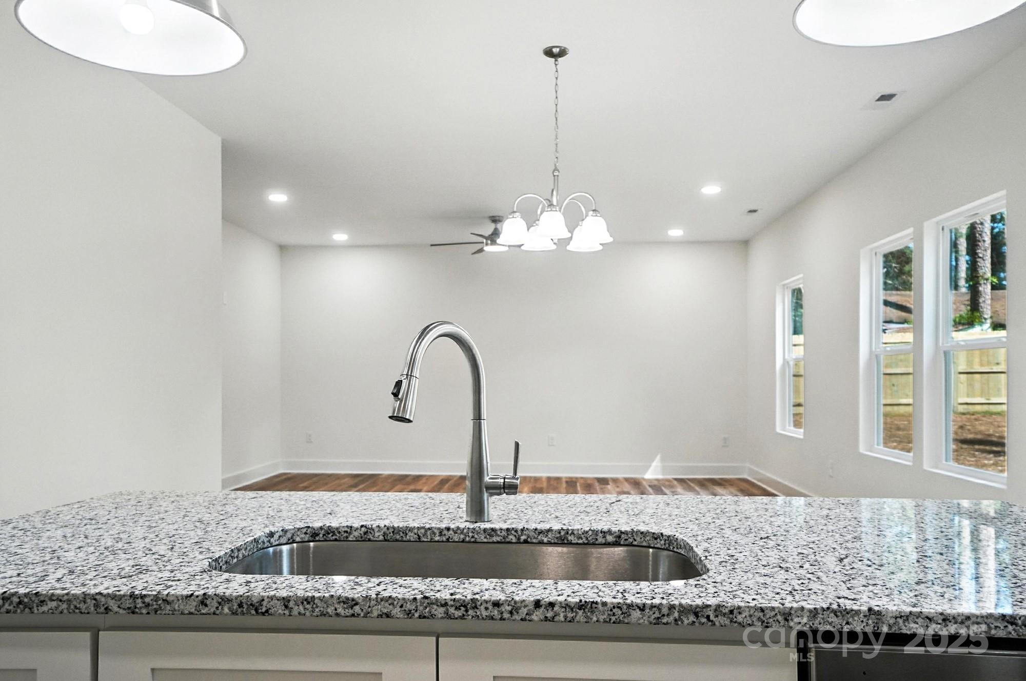 Kitchen sink and tall faucet set in a granite countertop, facing an empty dining area with windows.